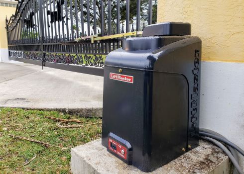 A technician mounts an electric gate opener motor to a concrete pad beside a black wrought iron sliding gate in Hallandale Beach.
